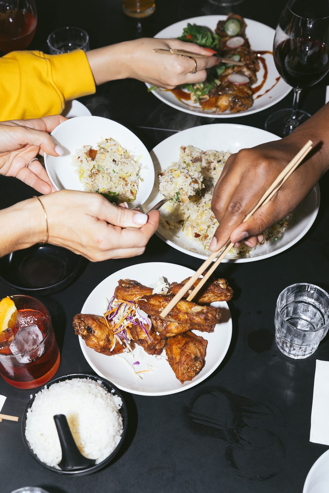 A shared banquet table at Happy Boy mid-service, hands reaching across plates of Chinese dishes and bowls of rice