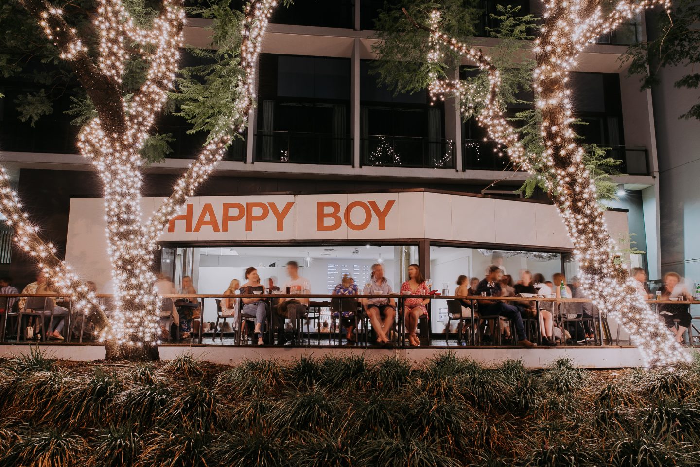 Happy Boy at night on East Street, the orange sign lit under fairy-lit trees with the dining room full behind glass