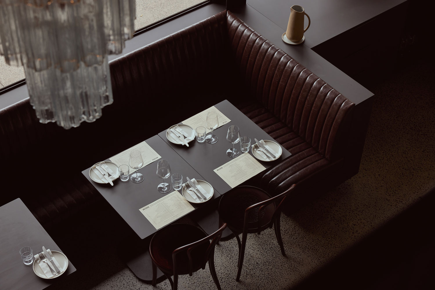 A curved leather booth at Petite set for dinner, beside the window and under a glass chandelier