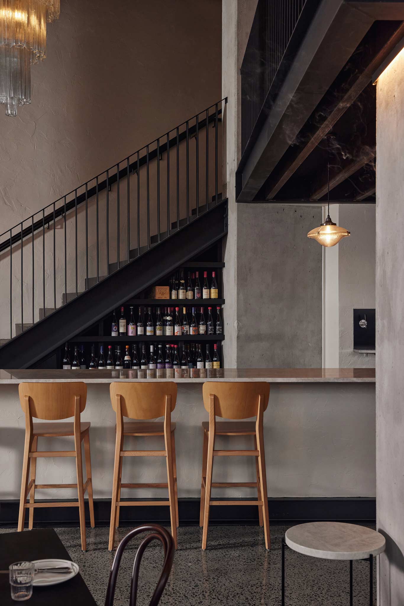 Petite's wine wall stacked with bottles beneath the mezzanine staircase, with stools at the marble bar
