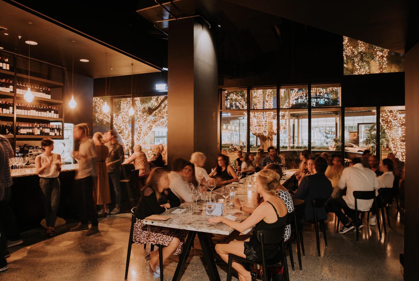 The main dining room at Snack Man with long marble tables and fairy-lit trees through the windows